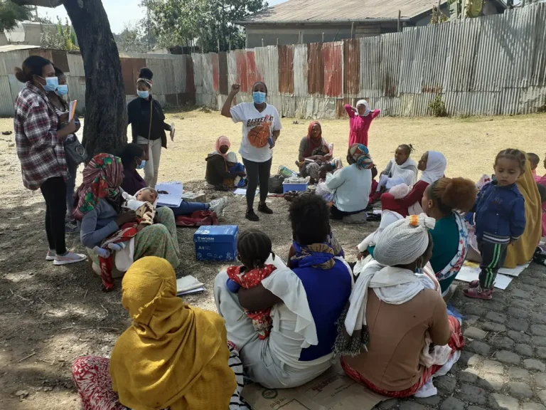 Facilitator leading a group of women and children seated outdoors.